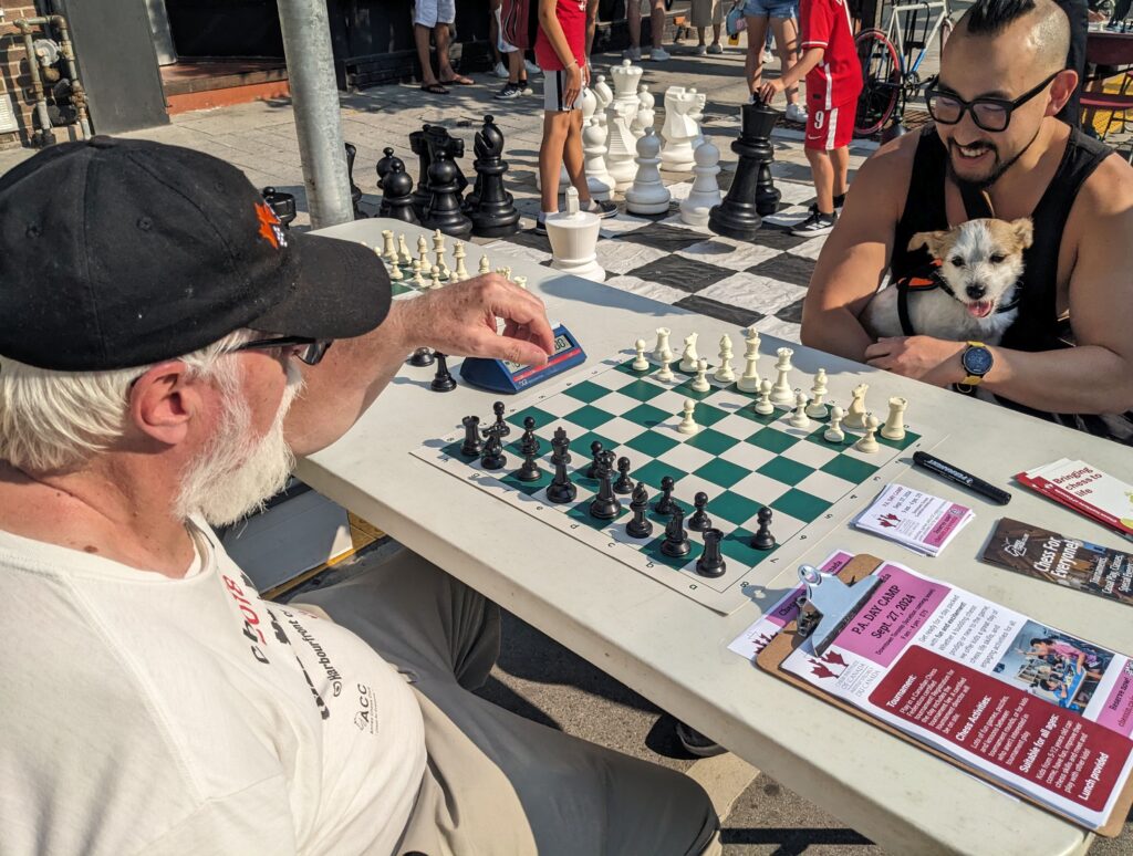 Two men play chess at outdoor event.